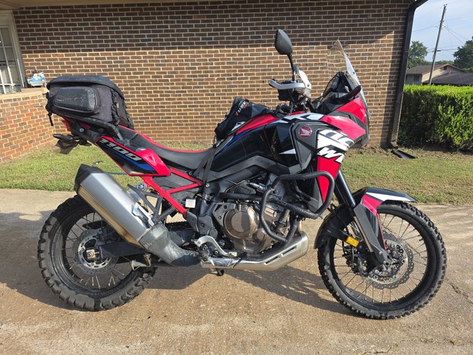 A red, black, and silver adventure motorcycle parked on a concrete driveway in front of a brick house, equipped with knobby tires, crash bars, and a rear luggage bag.