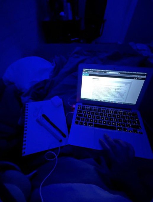A first-person perspective in a dark room illuminated by blue light, showing hands using the trackpad of a laptop displaying a document, with an open notebook and pen nearby.