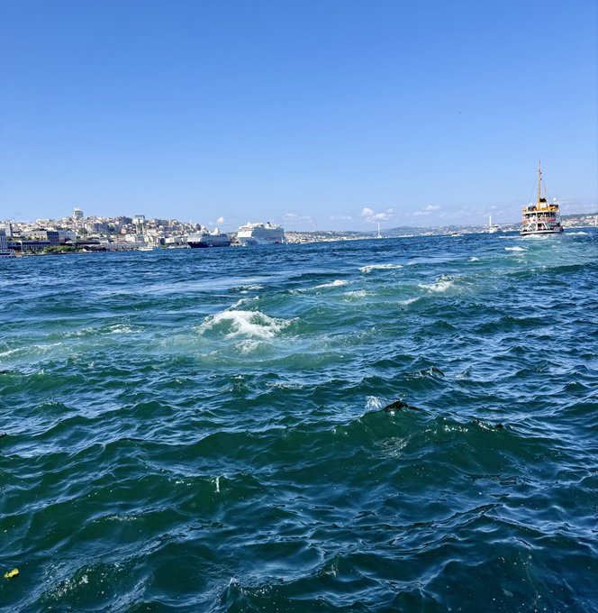 A view from the water showing blue choppy waves with a traditional ferry boat on the right, a large cruise ship, and a coastal city skyline in the distance under a clear blue sky.