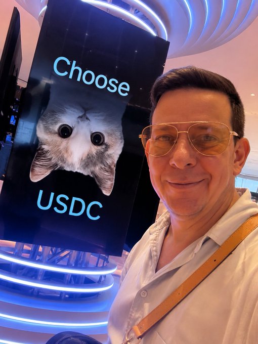 In the image, a man wearing sunglasses and a white shirt stands next to an advertisement for USDC. The advertisement features a cat with its face upside down, accompanied by the text "Choose USDC" written below it. The man appears to be posing in front of this unique advertisement, possibly as part of a promotional campaign or event related to USDC.