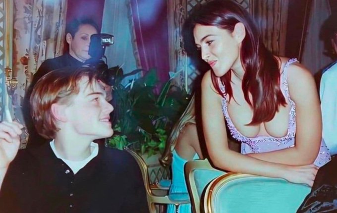 A young Leonardo DiCaprio smiles up at a woman in a pink top at what appears to be a glamorous event, with a photographer visible in the background. The image shows the kind of candid celebrity moment that someone asked Grok to recreate in higher resolution.