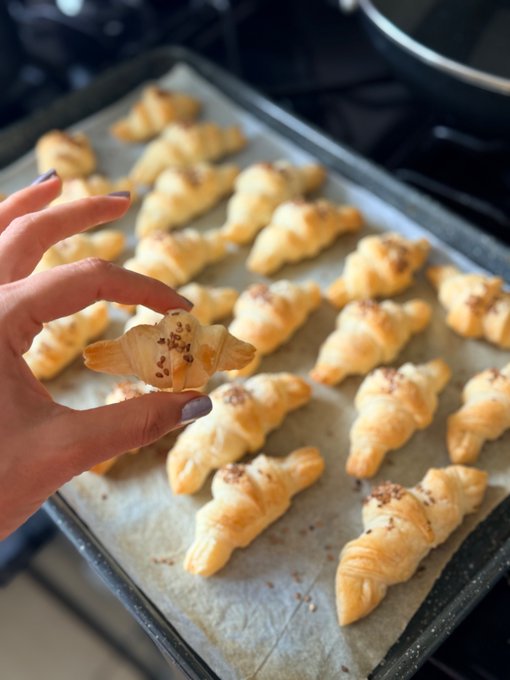 A hand holding up a small, freshly baked pastry above a baking sheet filled with many similar golden-brown pastries on a stovetop.