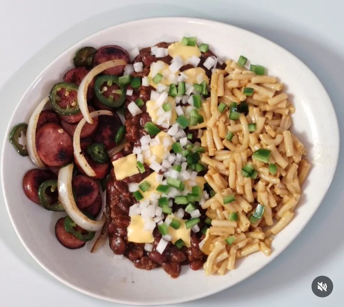A white plate filled with a meal consisting of chili with beans, yellow cheese, diced onions, and green onions, served alongside sliced sausage, onions, and jalapeños on one side, and macaroni on the other.