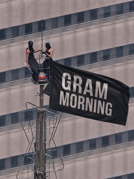 Two figures dressed in black stand atop a high antenna structure, unfurling a large black flag emblazoned with "GRAM MORNING" in white letters against the backdrop of a tall building. The banner's play on "Good Morning" pairs with the "GM" post, delivering a literal visual wake-up greeting to viewers below.