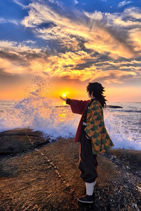 A cosplayer dressed as a character (appearing to be Giyu Tomioka from Demon Slayer) stands on a rocky shore at sunset, holding out their hand as if grasping the setting sun over the ocean, with waves crashing against the rocks.