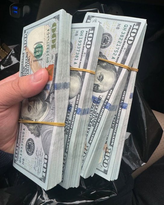 A hand holding several stacks of $100 US bills bound with rubber bands, appearing to total a large sum of cash.