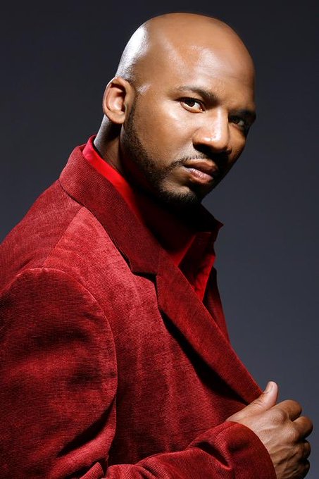A professional portrait of a bald Black man wearing a dark red velvet blazer over a red turtleneck, posed against a dark gray background.