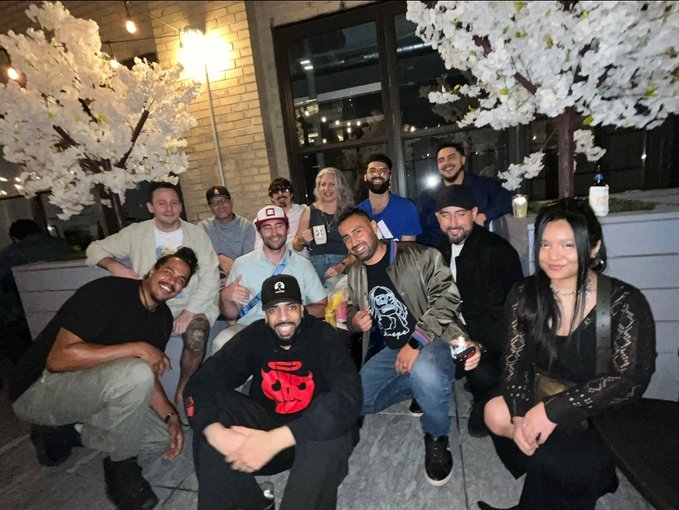 A group of nine people smiling and posing together at a nighttime gathering, with one man wearing a black t-shirt featuring a red seal/emblem design, surrounded by decorative white cherry blossom trees outside a building entrance.