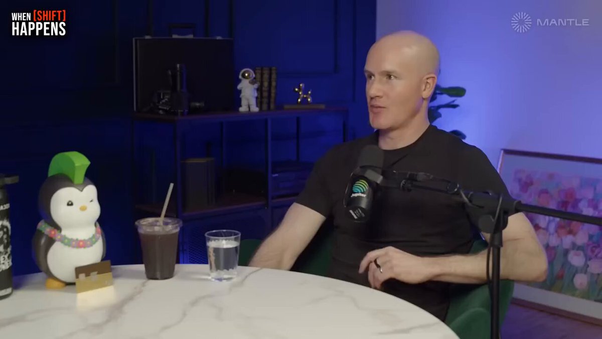 The image features a bald man sitting at a table with a microphone, likely participating in an interview or podcast recording session. The man appears to be wearing a black shirt and has a cup of coffee placed next to him on the table. There are two other cups visible in the scene - one near the bottom left corner and another closer to the center right side of the image.

The background includes a TV screen, suggesting that this might be an interview or discussion taking place at home or in a studio setting. The man's baldness is noticeable, making him stand out against the backdrop of the room.