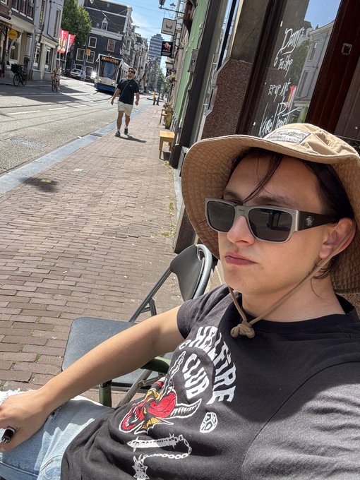 A selfie of a person wearing a tan bucket hat, dark sunglasses, and a black graphic t-shirt, sitting on a chair on a sunny city street with a tram and pedestrians in the background.