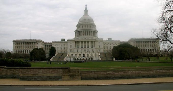 A view of the United States Capitol Building in Washington, D.C., taken from across a street with bare trees and a green lawn in the foreground under an overcast sky.