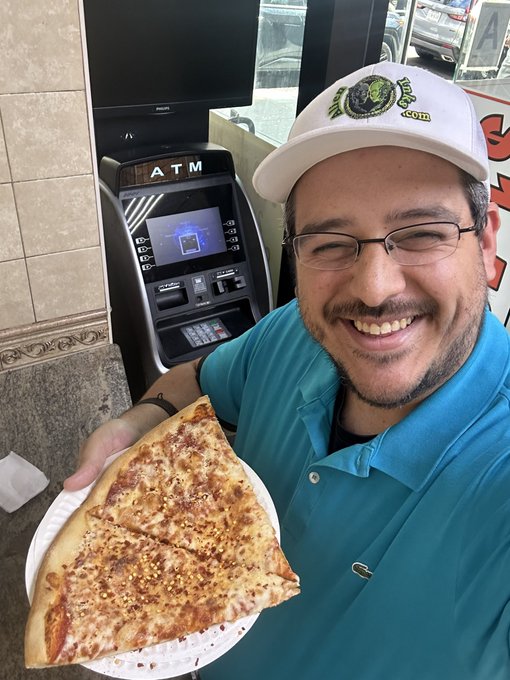 A smiling man wearing glasses, a teal polo shirt, and a white cap holds a paper plate with a large slice of pizza in front of an ATM machine.