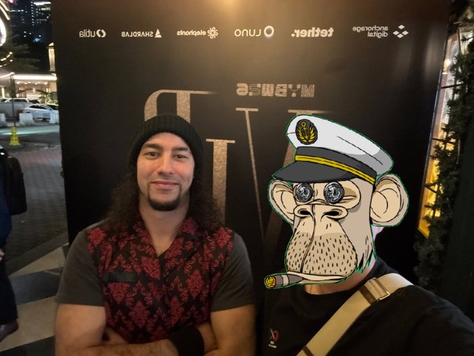 A man with long hair and a beanie stands next to another person whose face is replaced with an 'Ape Strong Together' cartoon ape wearing a sailor hat, posing in front of a black sponsor backdrop.