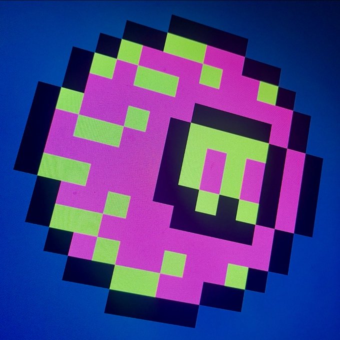 A pixel art-style circular graphic featuring pink, green, and black blocks forming an abstract character or logo against a deep blue background.