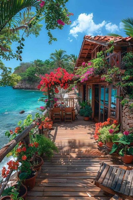 A vibrant tropical scene featuring a wooden deck and rustic house surrounded by blooming pink and red bougainvillea flowers, overlooking turquoise ocean waters with palm trees under a bright blue sky.