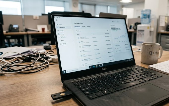 A Dell laptop on a wooden desk displays a financial transactions dashboard showing a value of $980,000.00, with a mug and tangled cables nearby.
