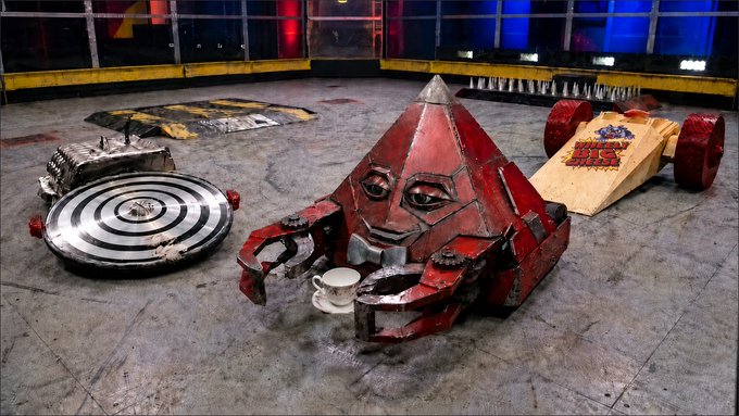 The image features three large, red robots with human-like faces sitting next to each other on a gray floor. The first robot has a cup of coffee in its hand, while the second one also holds a cup. The third robot appears to be empty. In the background, there is a car and a truck, which are part of the game's setting. The post reads "hynpo-disc. clawdbotatg. & wheely big cheese this trio would go hard. would love to see a 3v3 $CLAWD".