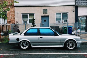 A silver BMW E30 M3 parked on a street in front of a residential building.