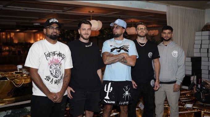 Five men pose together indoors, with the central figure wearing a light blue cap and t-shirt, flanked by others in casual streetwear including a white graphic tee and a gray hoodie.