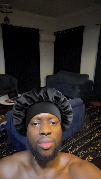 A shirtless man wearing a black satin bonnet taking a selfie in a dimly lit living room with dark curtains and a sofa.