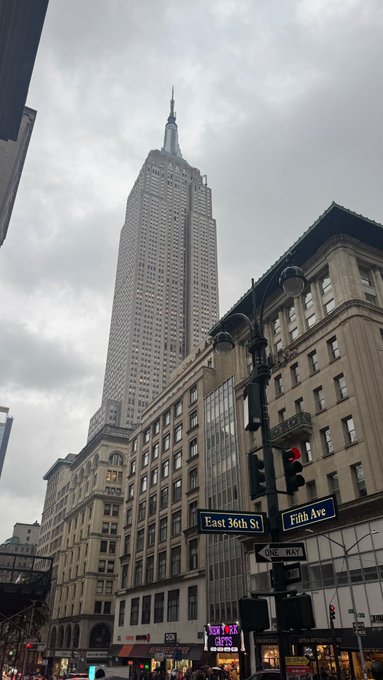 A street-level view shows the Empire State Building towering under a cloudy sky, with street signs for 'East 36th St' and 'Fifth Ave' visible at the intersection.