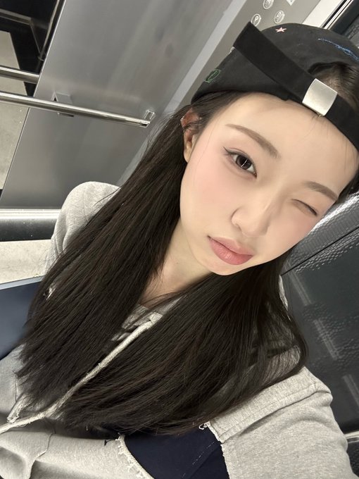 A young woman wearing a backwards black cap and a grey zip-up hoodie winks at the camera in a close-up selfie taken inside what appears to be public transit.