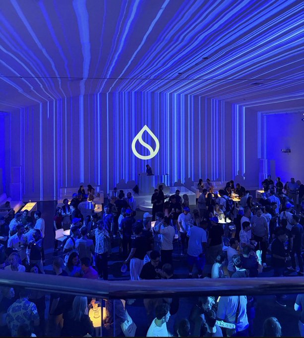 A large crowd gathers in a room illuminated by striking blue radiating light patterns, with a glowing water drop logo prominently displayed at the center of the stage, representing the Sui blockchain community event. The vibrant atmosphere and engaged attendees visually reinforce the post's message about Sui's strong, united community.