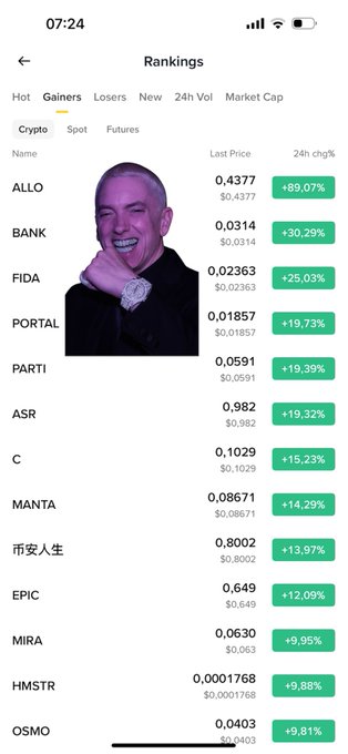 The image shows a phone screen displaying a list of cryptocurrency names, along with their respective market caps and rankings. The topmost name among these is "ALLO", which appears at the very top of the list. There are several other names on the list, including "HOST" and "MANT". The phone screen also displays the text "uptrend chào các tỷ phú tương lai ALLO mà ăn nằm ở gainers tần 2 tuần thì ... make ALLO great như LAB". The phone screen is positioned on a table, and the focus of the image is on this list.