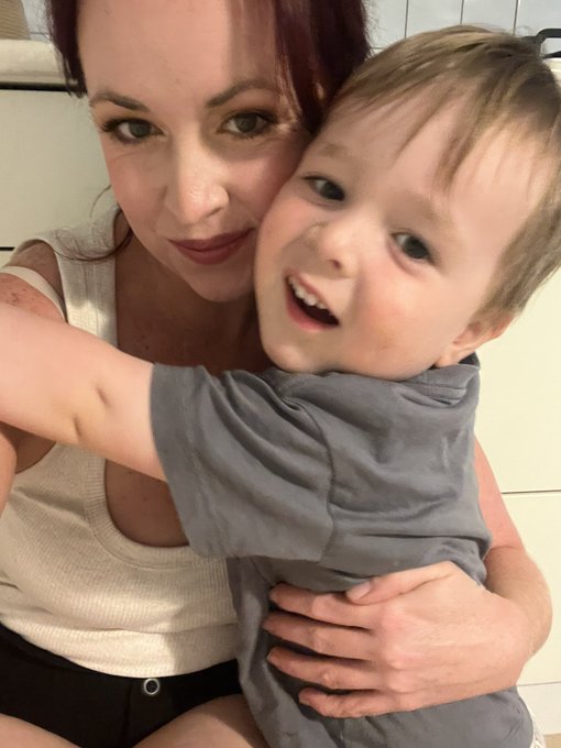 A close-up selfie of a smiling woman with red hair holding a young boy in a gray t-shirt who is grinning happily at the camera.