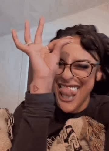 A selfie of a smiling woman with glasses making an 'OK' hand gesture over her eye, wearing a dark top and a patterned blanket.