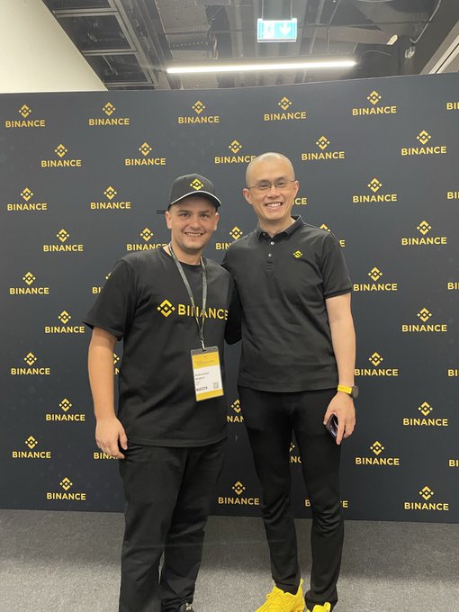Two men wearing black shirts with the yellow Binance logo pose together in front of a Binance-branded backdrop at what appears to be a corporate event.