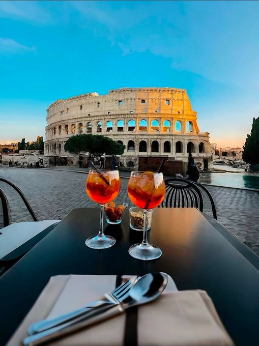 Two glasses of aperol spritz on a table with the Colosseum in Rome visible in the background at dusk.