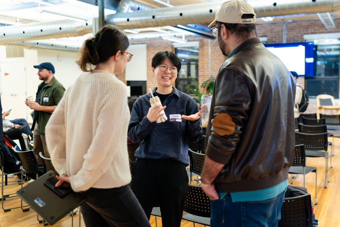 Three people converse in an open, modern indoor space with exposed brick and industrial ceilings, with one person holding a clear bottle.