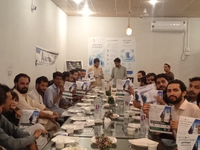 A group of men gather around a long banquet table in Pakistan, holding up pamphlets titled "AILYTIS & ODBIT" featuring images of a water pitcher, with matching AILYTIS banners displayed on the walls behind them during the community event organized by Usman.