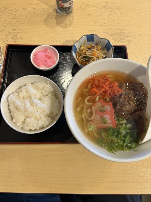 A Japanese meal set on a black tray features a bowl of white rice, a bowl of udon noodle soup with beef and vegetables, and small side dishes including pink pickled ginger and kinpira-style vegetables.