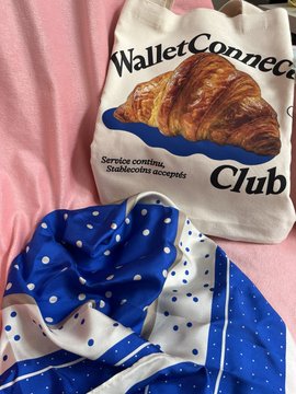 A canvas tote bag reads 'WalletConnect Club' with a croissant illustration and the tagline 'Service continue. Stubbornly accept', resting near a blue and white polka-dot fabric.