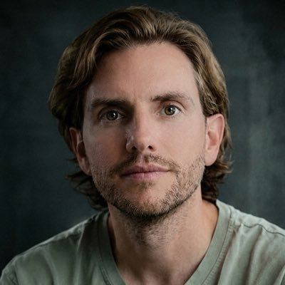 A portrait of a man with shoulder-length brown hair and a short beard, wearing a light-colored t-shirt, shown against a dark background. The image accompanies news about Chainlink's Chris Barrett discussing the CLARITY Act's anticipated advancement in a US Senate procedural vote on digital asset legislation.