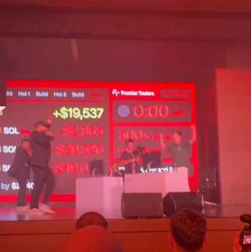 A dimly lit photo of people on a stage at an event with red screens displaying 'Primitive Traders' with '+19,500' prizes.