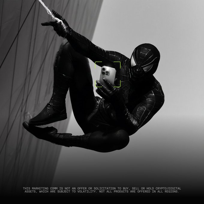 The image depicts Spider-Man in his black symbiote suit, suspended in mid-air by a web, while examining a smartphone with a viewfinder overlay—suggesting a trading or scanning moment. The accompanying text "A brand new trade starts now. Are you ready?" frames the scene as a teaser for a new crypto trading opportunity, with a disclaimer at the bottom noting that the marketing communication is not an offer or solicitation to buy, sell, or hold digital assets, which are subject to volatility and may not be offered in all regions.