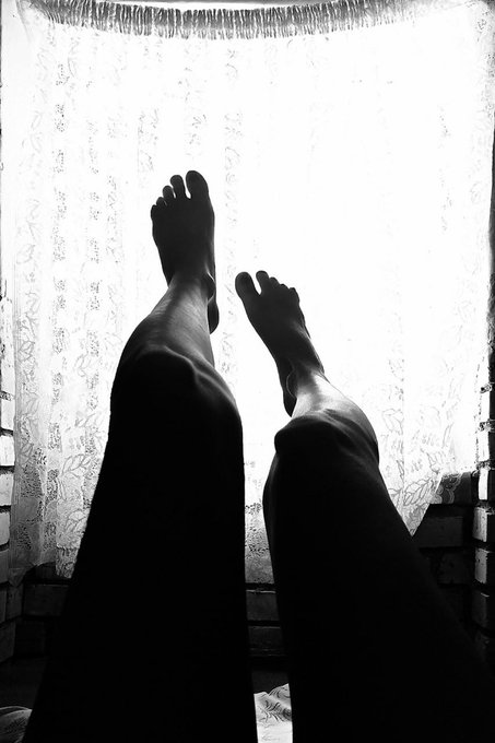 A black and white silhouette photograph showing a person's legs and bare feet raised up against a bright window with sheer lace curtains.
