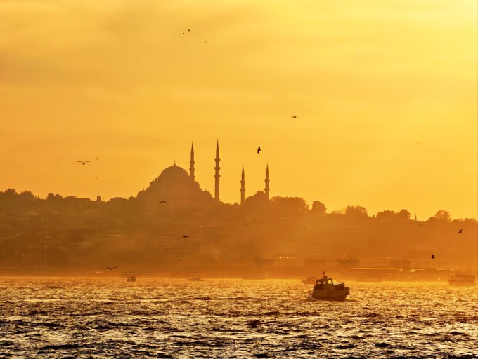 A golden-orange sunset silhouettes a waterfront mosque with domes and minarets, while birds and small boats appear over shimmering water.