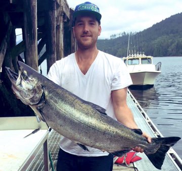 A man in a white t-shirt and 'IRON' cap proudly holding a large salmon he caught, standing on a dock with a boat and forested hills in the background.