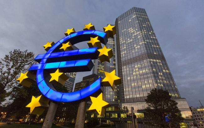 A large illuminated Euro sign sculpture surrounded by yellow stars stands in front of a modern glass skyscraper at dusk.