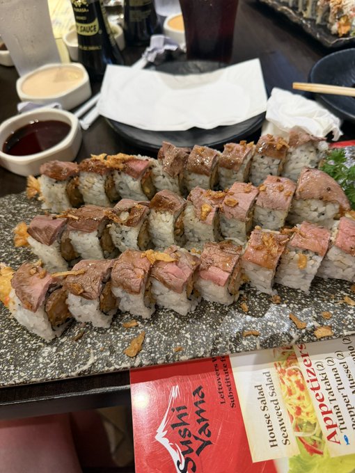 A large platter of sushi rolls topped with seared beef from Sushi Yama, accompanied by dipping sauces, chopsticks, and an appetizer menu.