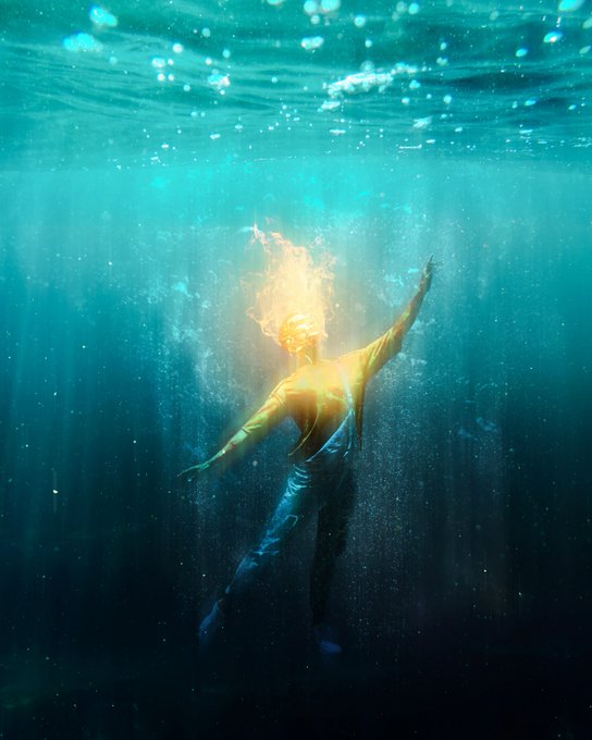 A person appears submerged underwater with their head engulfed in bright, glowing flames, arms outstretched in the blue depths.
