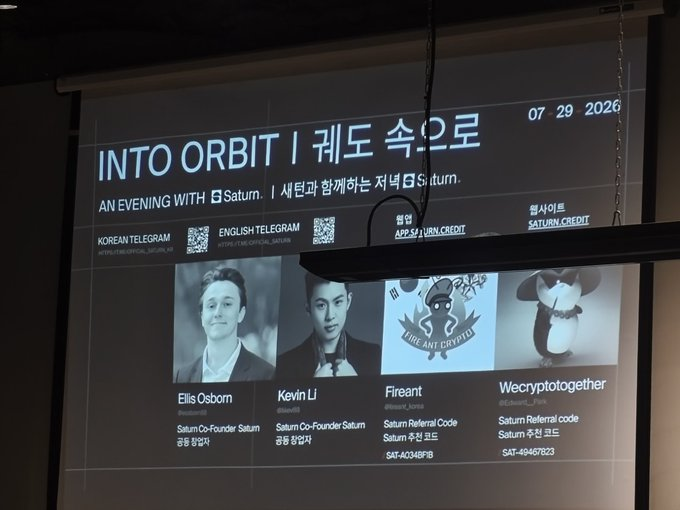 A photograph of a projection screen at an event titled 'INTO ORBIT | 궤도로 속으로' (dated 07/29/2026) showcasing four speakers/sponsors: Ellis Osborn (Saturn Co-Founder), Kevin Li (Saturn Co-Founder), Fireant (Saturn Referral Code), and Wecryptogether (Saturn Referral Code), with QR codes for Korean and English Telegram channels.
