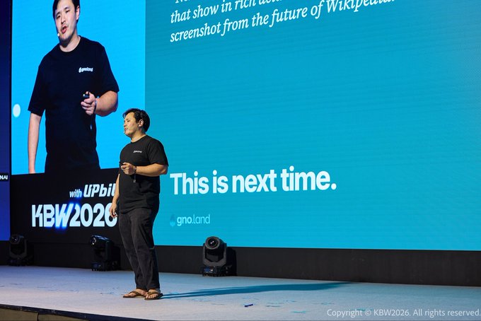 A man in a black t-shirt and sandals presenting on stage at KBW2026, with the text "This is next time." and the gno.land logo displayed on the large screen behind him.