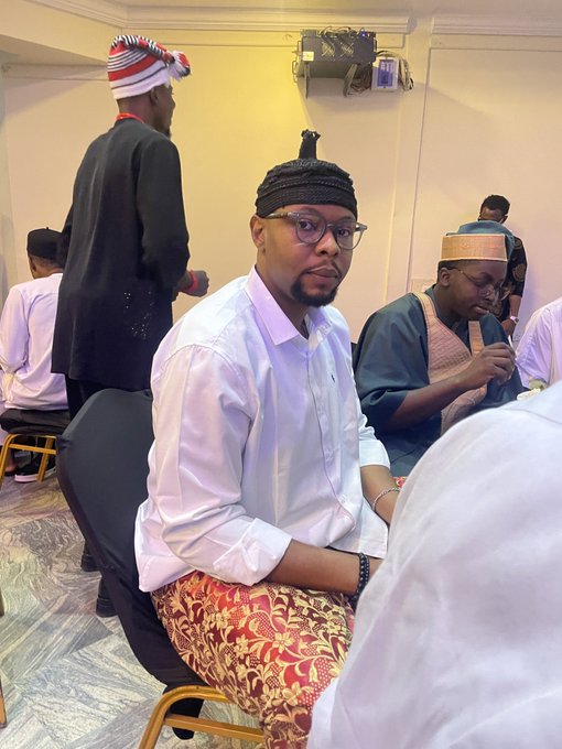 A young man wearing glasses, a traditional cap, and a patterned wrapper sits among other attendees at the Solana Summit, appearing to pose for the photo in a formal cultural setting.