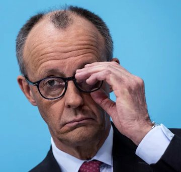 A man in a dark suit with a red tie adjusts his round black glasses, looking thoughtfully at the camera against a blue background.