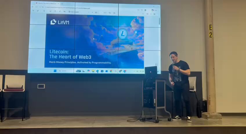The image shows a man standing behind a podium, giving a presentation or speech at a conference. The man appears to be wearing a black shirt and has his hands up as if he's emphasizing something during his talk. The backdrop of the scene is a large screen displaying information related to Litecoin, possibly about its history or future implications in the world of cryptocurrency.